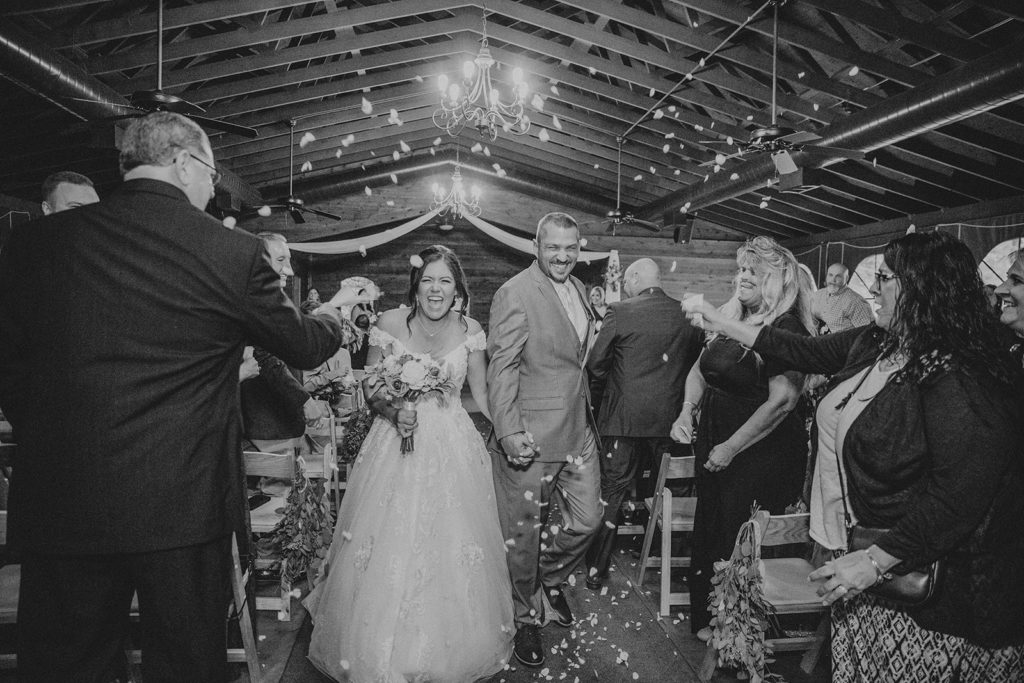 Couple just married coming back down the aisle laughing as flower petals are thrown over top of them at Bluebridge Event Center in Grawn Michigan. cut your guest list