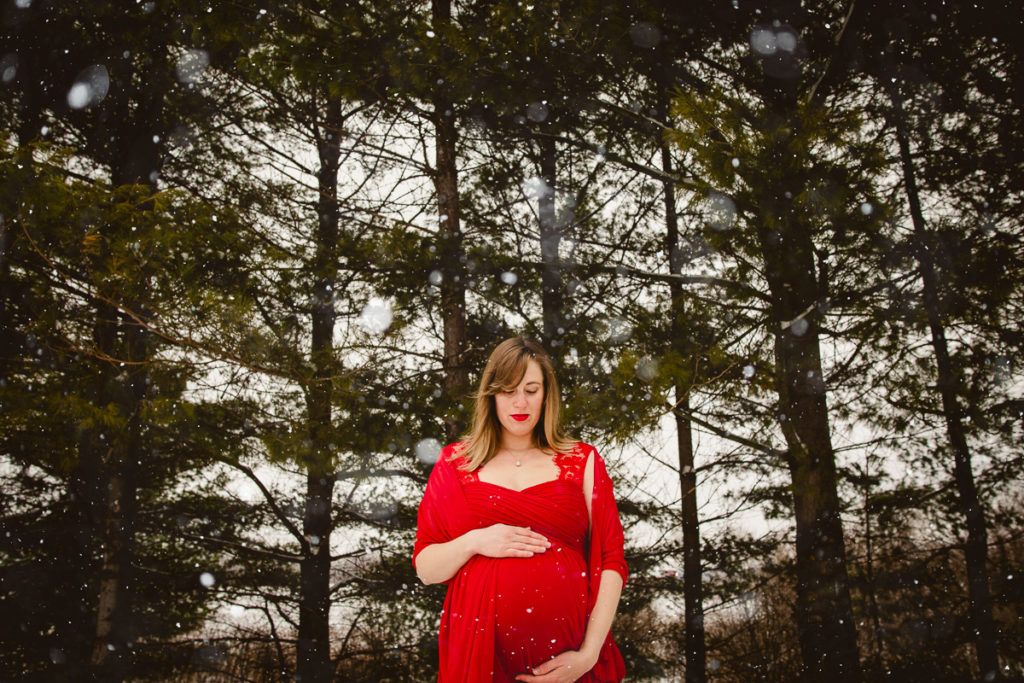 A pregnant woman, in a red dress, looking down her dress in front of the pine tress, while it snows. Maternity session outfits dos & don'ts