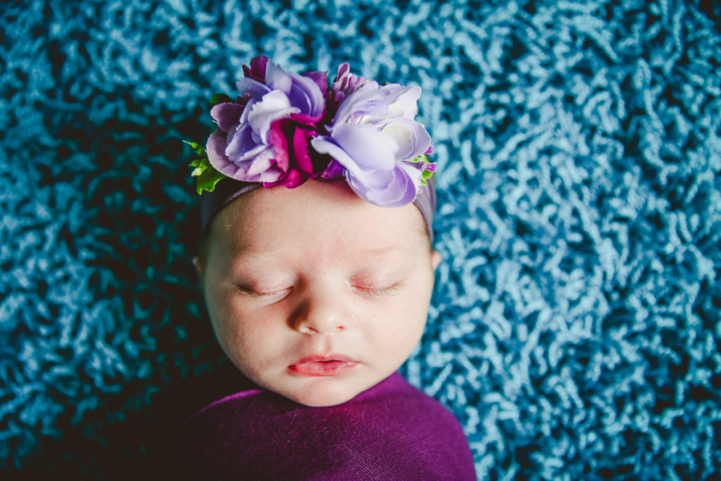 Newborn laying on the carpet wrapped in purple swaddle. 