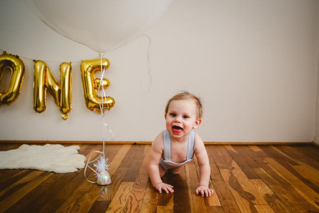 A little boy crawling on the floor in front of gold balloon one for one year. Milestone session planning guide.