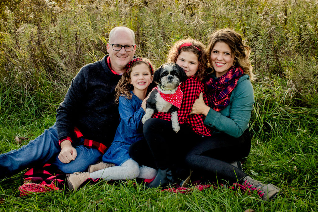 Family sitting on the ground on a blanket in holiday colors with their little dog all looking at the camera. 5 tips to include your dog. 