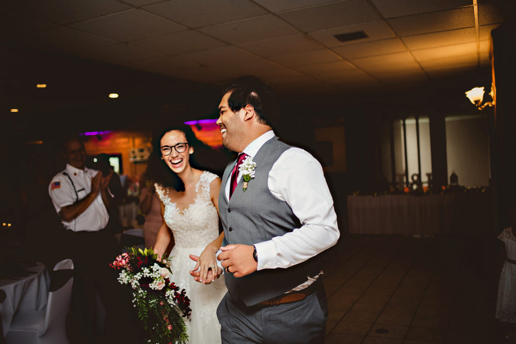 A couple laughing running in to their wedding reception after being announced. Groom is looking at the bride while the bride is laughing while looking at the camera. 