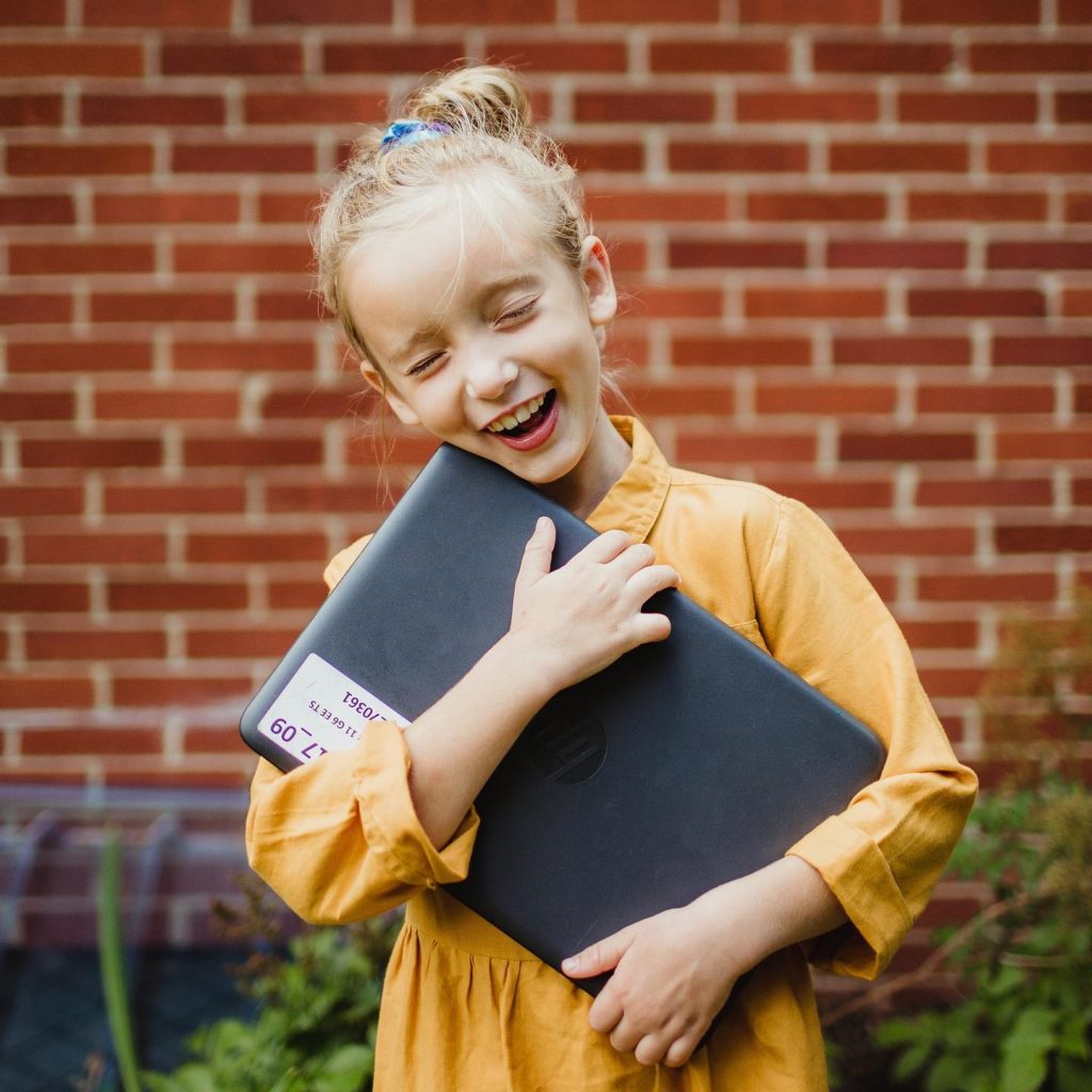 A blonde little girl wearing a yellow dress laughing while hugging a Chrome Book