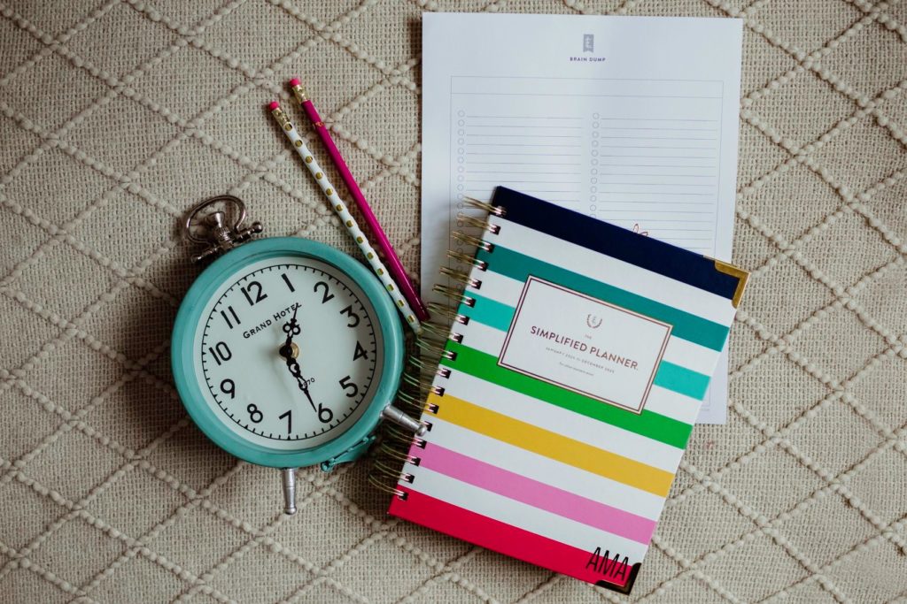 A teal clock set to 1:30pm laying next to some pencils a Happy Stripe Emily Ley Simplified planner with pencils & paper