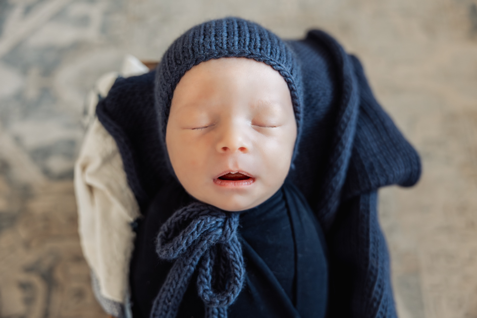 Newborn baby sleeping peacefully in a dark blue knit bonnet and swaddle during an in-home session